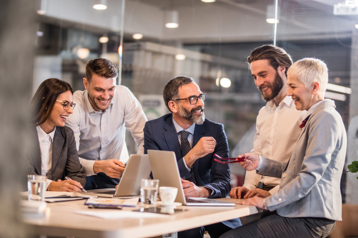 A group of five people in business attire are sitting and standing around a table, engaged in a discussion about sales pipeline management in a modern office setting with laptops and documents.