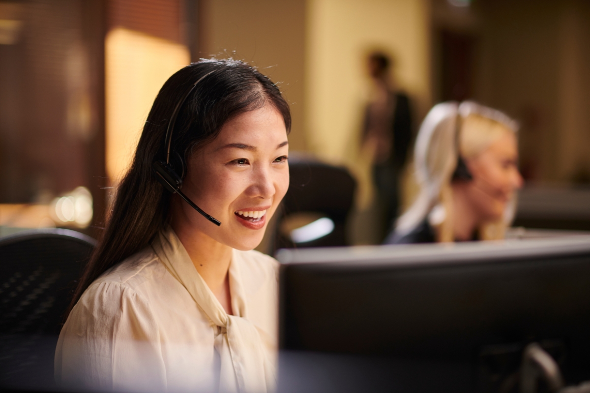 A woman with a headset is smiling while managing the sales pipeline on her computer in an office. Another person with a headset is visible in the background.