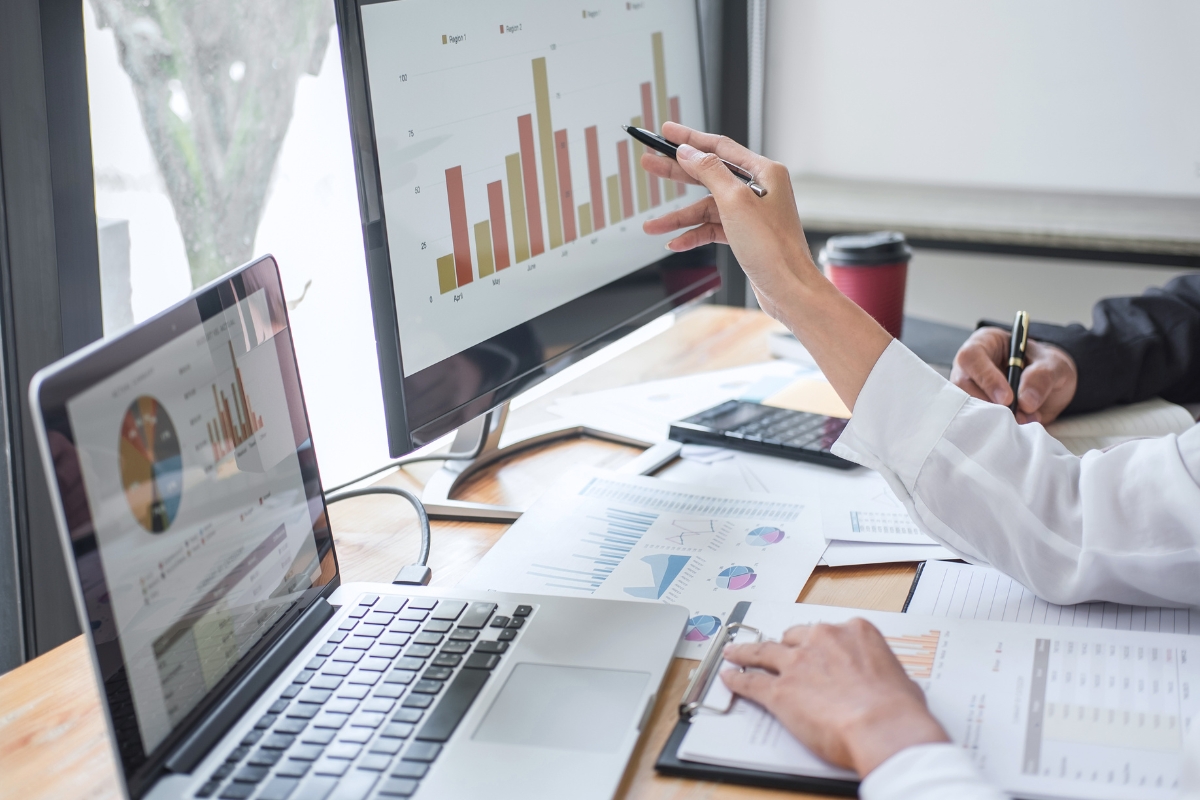 People working on laptops and examining printed charts and a bar graph on a monitor; one person is pointing at the screen with a pen, discussing sales pipeline management.