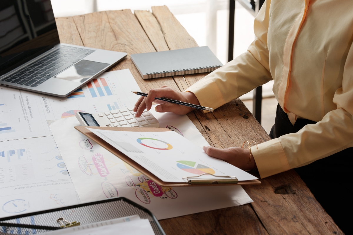 Person in a yellow shirt using a calculator and reviewing financial charts at a wooden desk with a laptop and notebook, focusing on sales pipeline management.