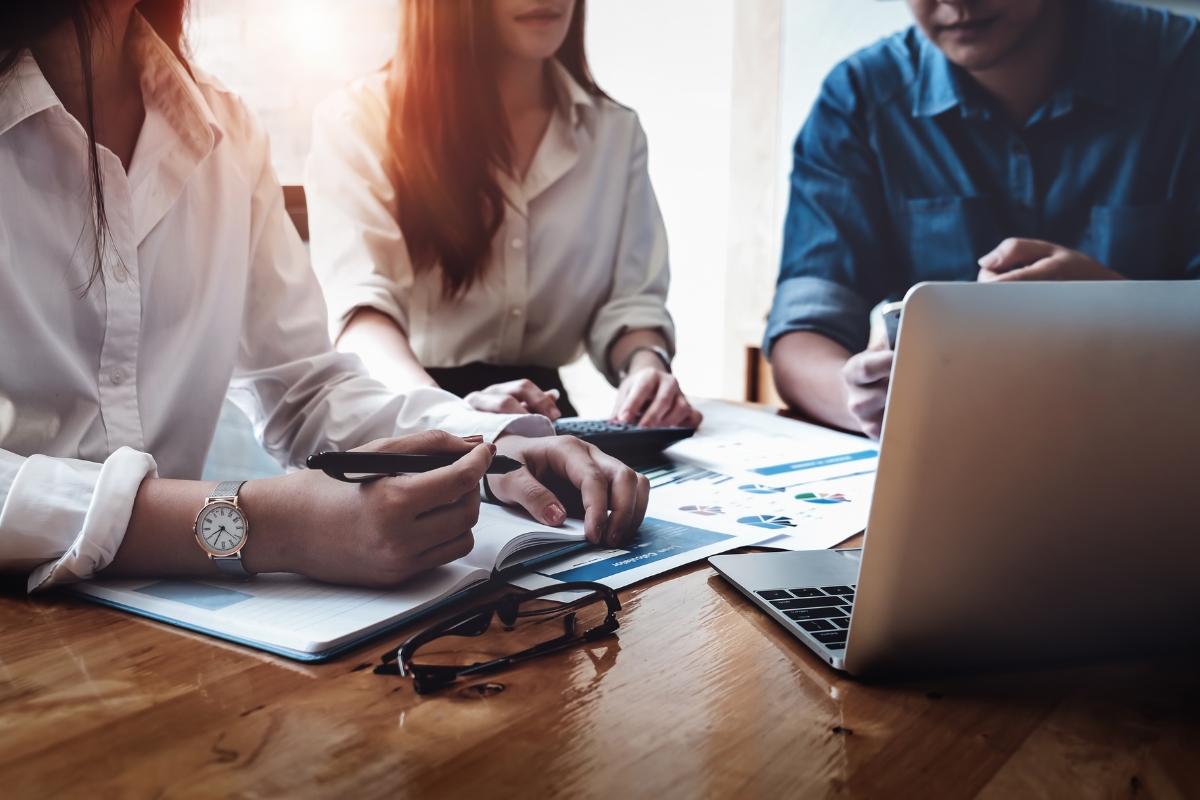 Three individuals collaborate at a wooden table, referencing documents and working on a laptop to enhance their sales process automation. A pair of glasses is placed on the table next to them.