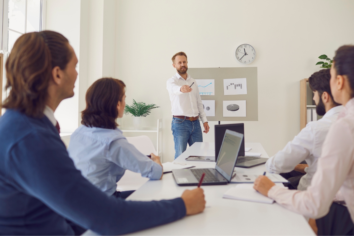 A man in a white shirt presents visuals on a board to four seated colleagues in a meeting room, discussing sales process automation. Laptops and papers are on the table, and a clock is on the wall.