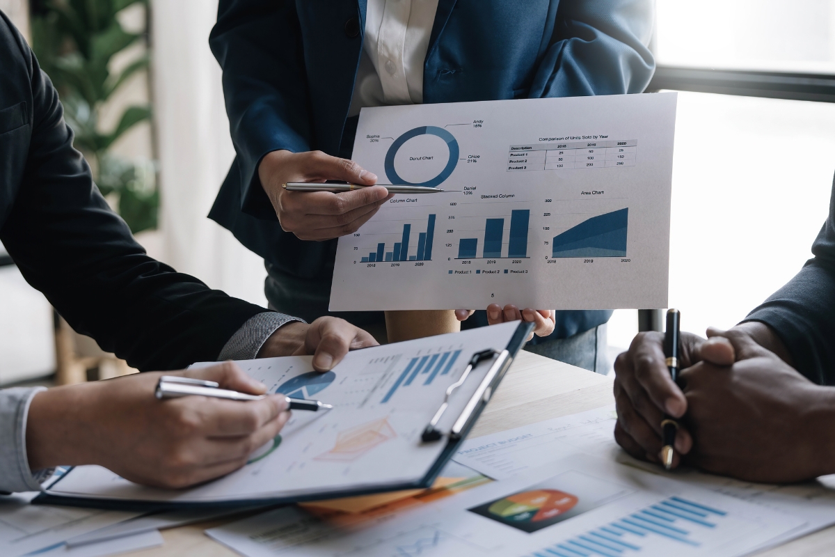 Three people discuss charts and graphs during a business meeting, with one person holding up a paper displaying various data visualizations related to sales process automation.