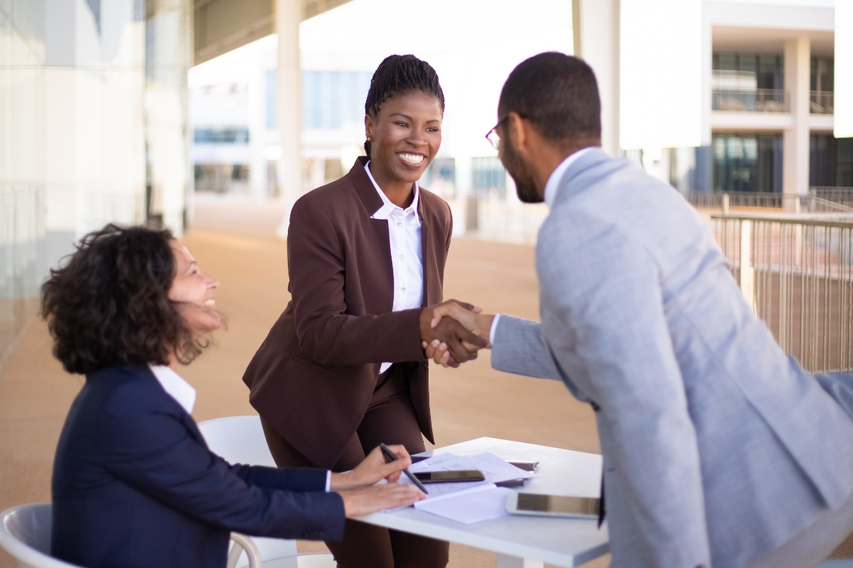 Three professionals having a meeting outdoors. A woman in a brown suit is shaking hands with a man in a light gray suit, while another woman in a dark blue suit takes notes at the table, discussing sales process automation.