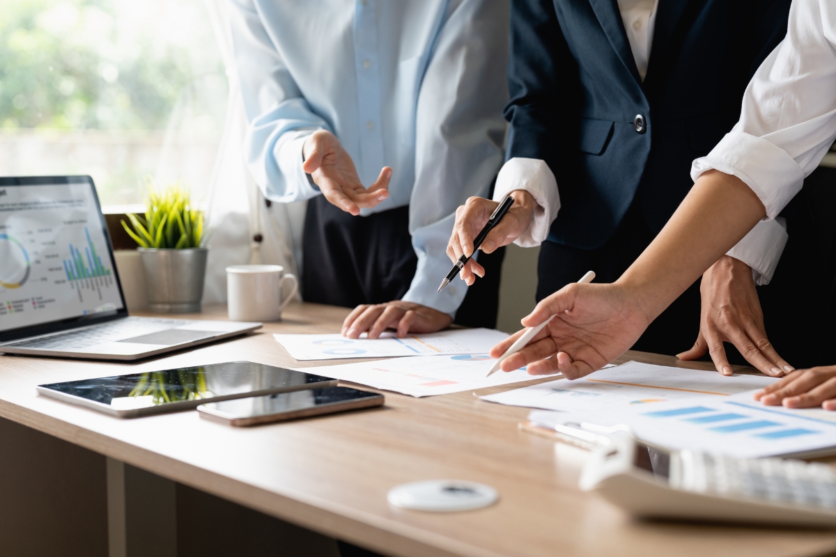 Three individuals in business attire are discussing paperwork with charts and graphs on a table, likely focusing on sales process automation. A laptop, tablet, and other office supplies are also on the table.