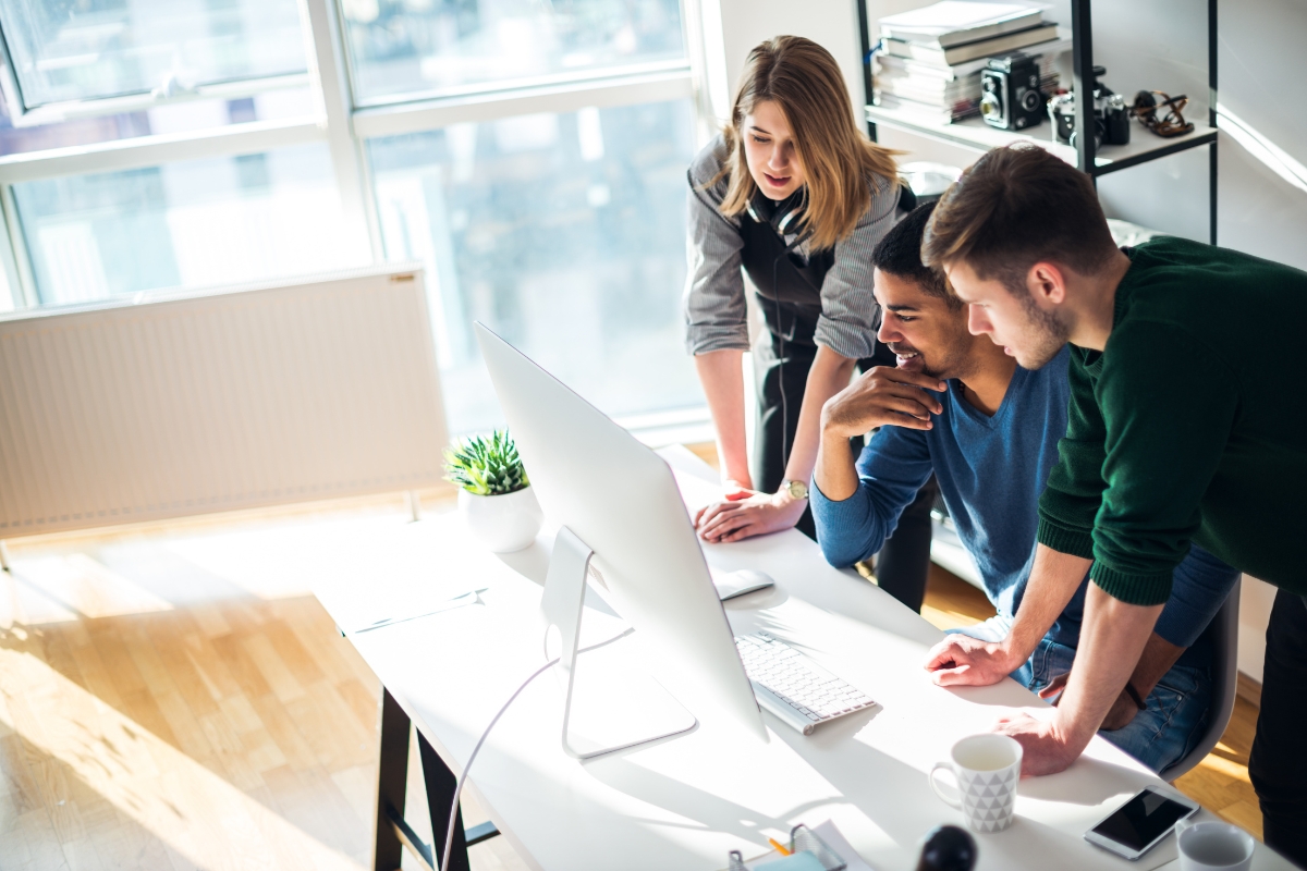 Three colleagues gathered around a desk look at a computer screen, working together on sales process automation in a bright office space. Various office supplies and a coffee mug are on the desk.