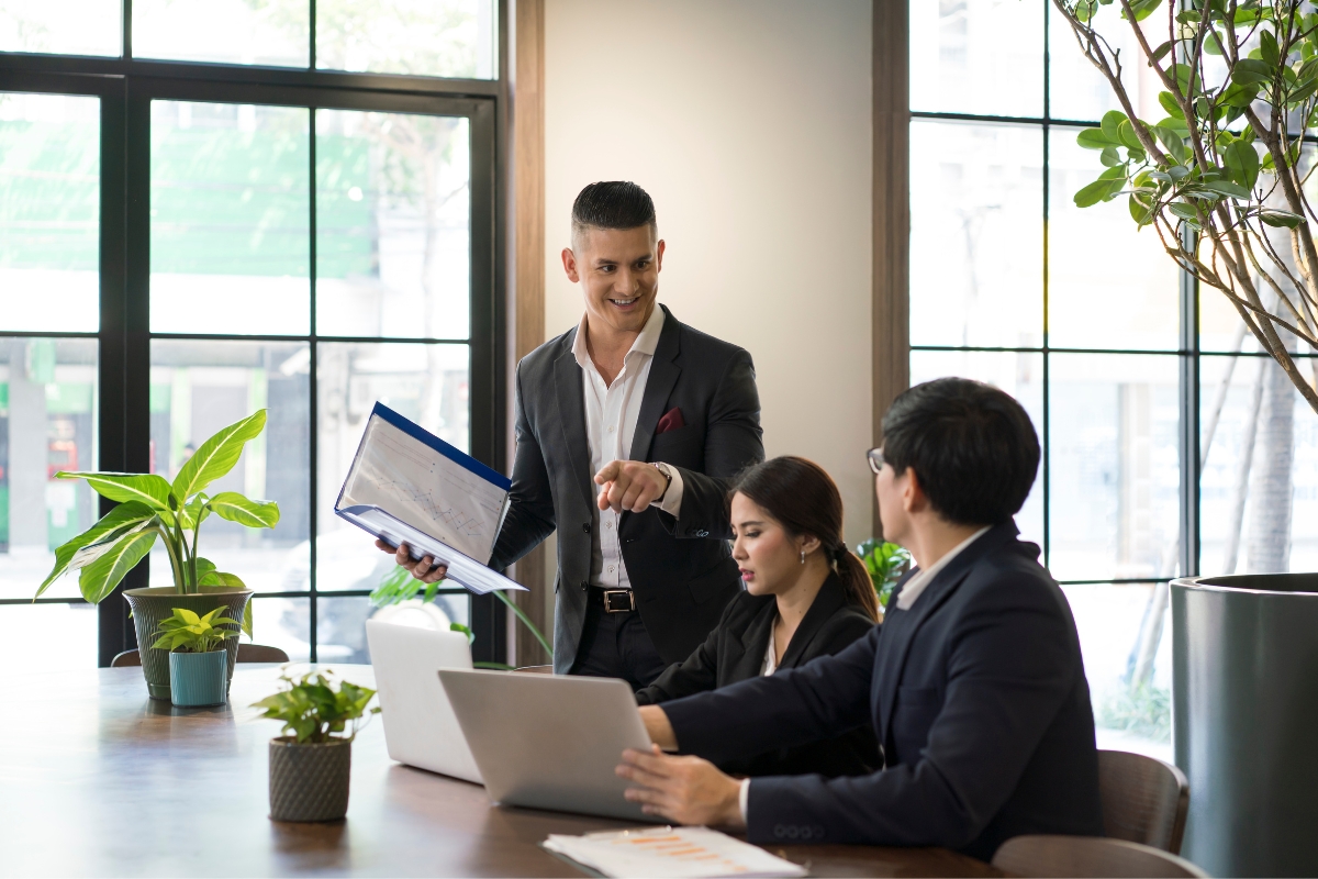 Three business professionals discuss sales process automation in a modern office with large windows and green plants. One man stands holding a document, while a woman and another man are seated with laptops.