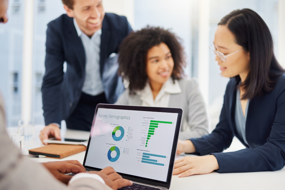 Four people in business attire are gathered around a laptop displaying survey demographics charts. They appear to be discussing the data shown on the screen, strategizing on how to transform customer service based on the insights revealed.