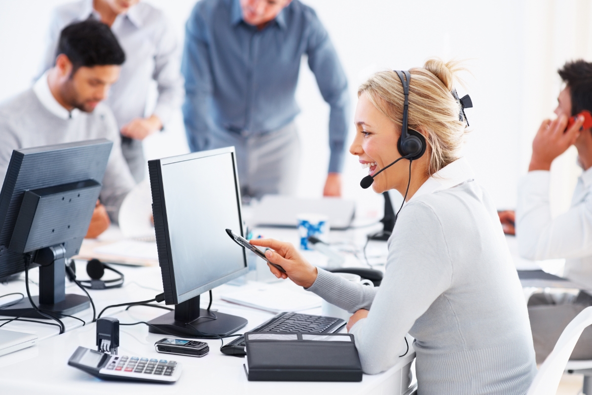 A woman wearing a headset works at a computer in an office, aiming to transform customer service, with three colleagues in the background.