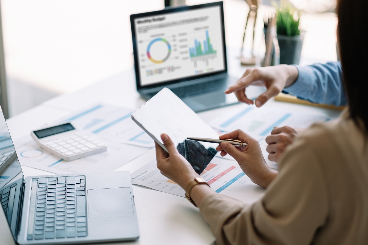 Two people reviewing graphs and charts on a tablet and laptop at a desk with financial documents, a calculator, and a plant, aiming to transform customer service.