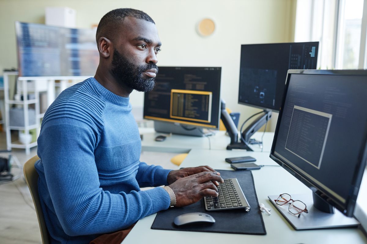 A man in a blue sweater types on a keyboard at a desk with multiple computer monitors displaying code, focused on optimizing workflow automation.