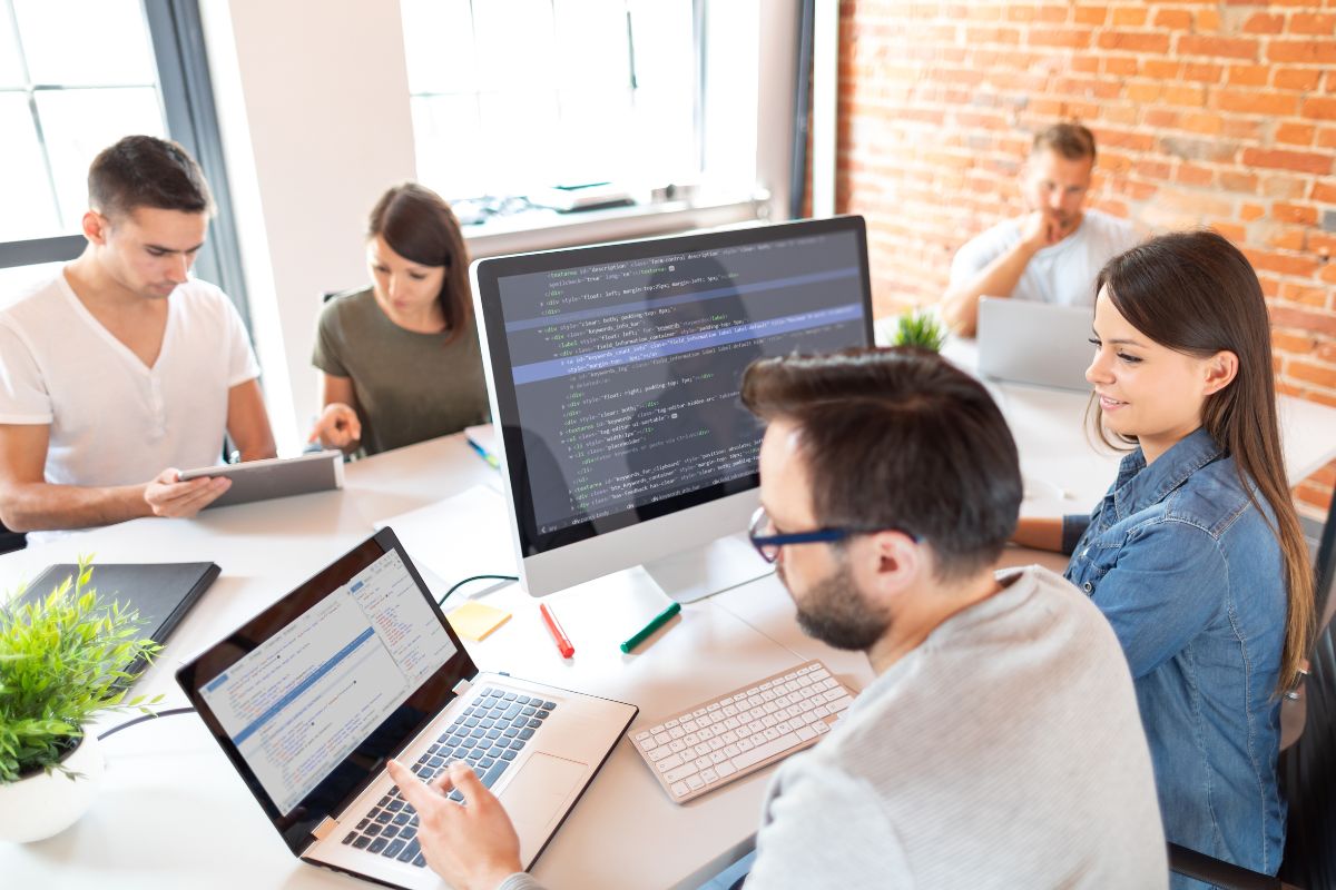 A group of five people is working at a table with laptops and desktop computers; two are looking at code on a monitor. The setting appears to be a modern office with brick walls and large windows, where they seem to be developing a CRM for business automation.