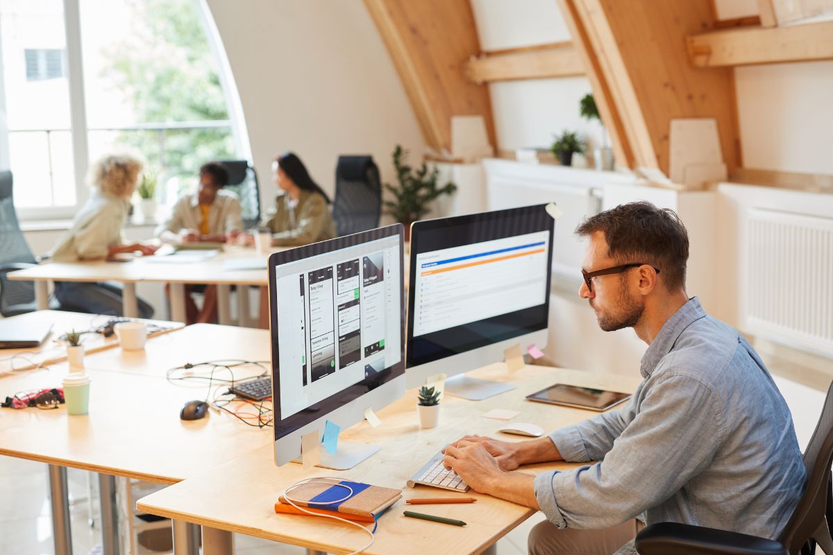 A person works at a desk with two monitors, seamlessly integrating workflow automation, while three people have a meeting in the background in a modern office with wooden beams and large windows.