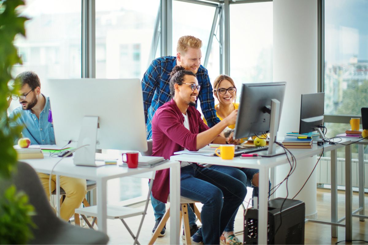 Three people are gathered around a computer screen in an office, discussing workflow automation with smiles. Another person is working at a separate desk. The office space has large windows and natural light.