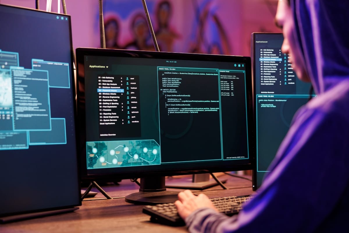 A person in a hoodie works at a computer surrounded by multiple screens displaying code, graphs, and cloud backup services in the dimly lit room.