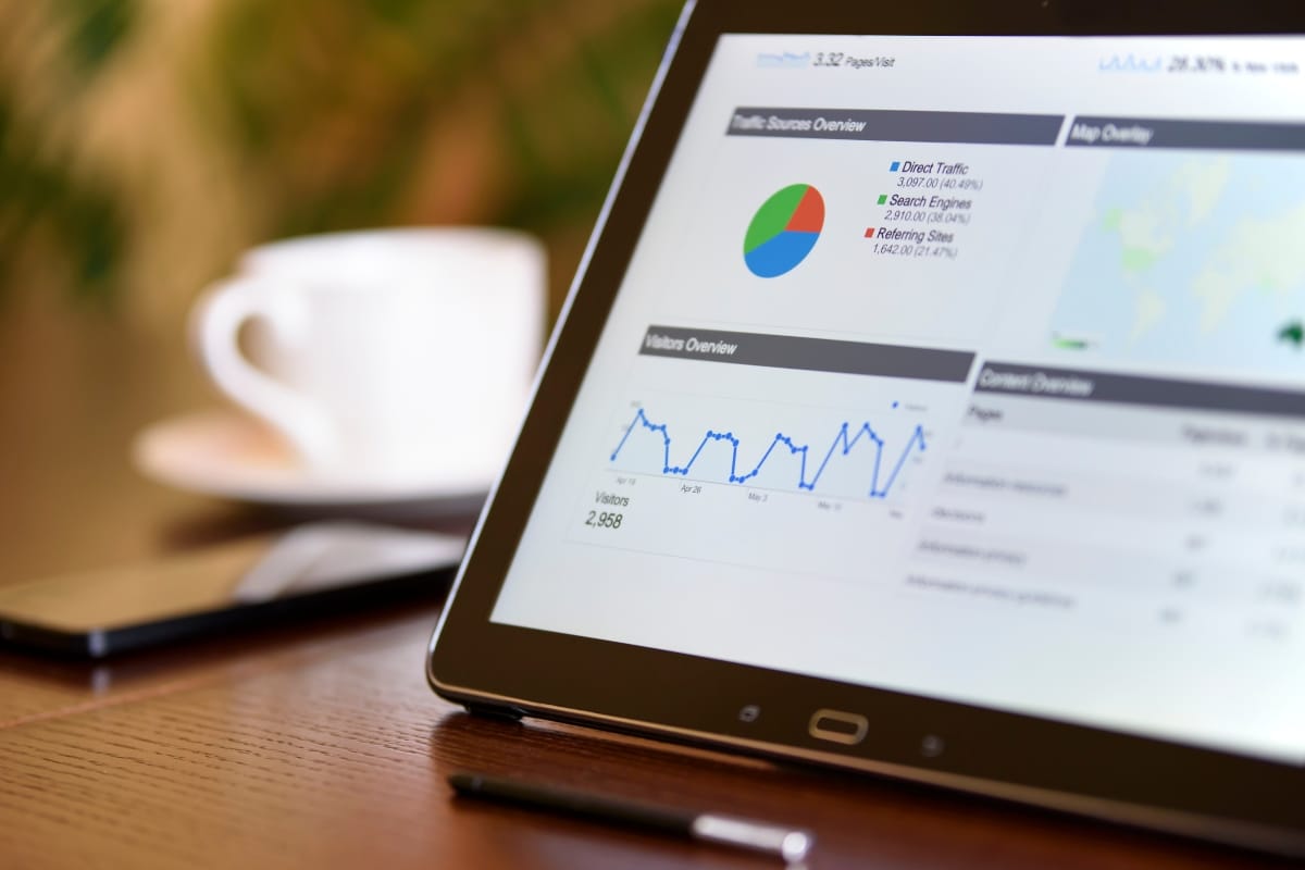 A tablet displaying digital analytics with charts and graphs sits on a wooden table, alongside a content calendar planning sheet. Nearby, there is a smartphone, a pen, and a white coffee cup.