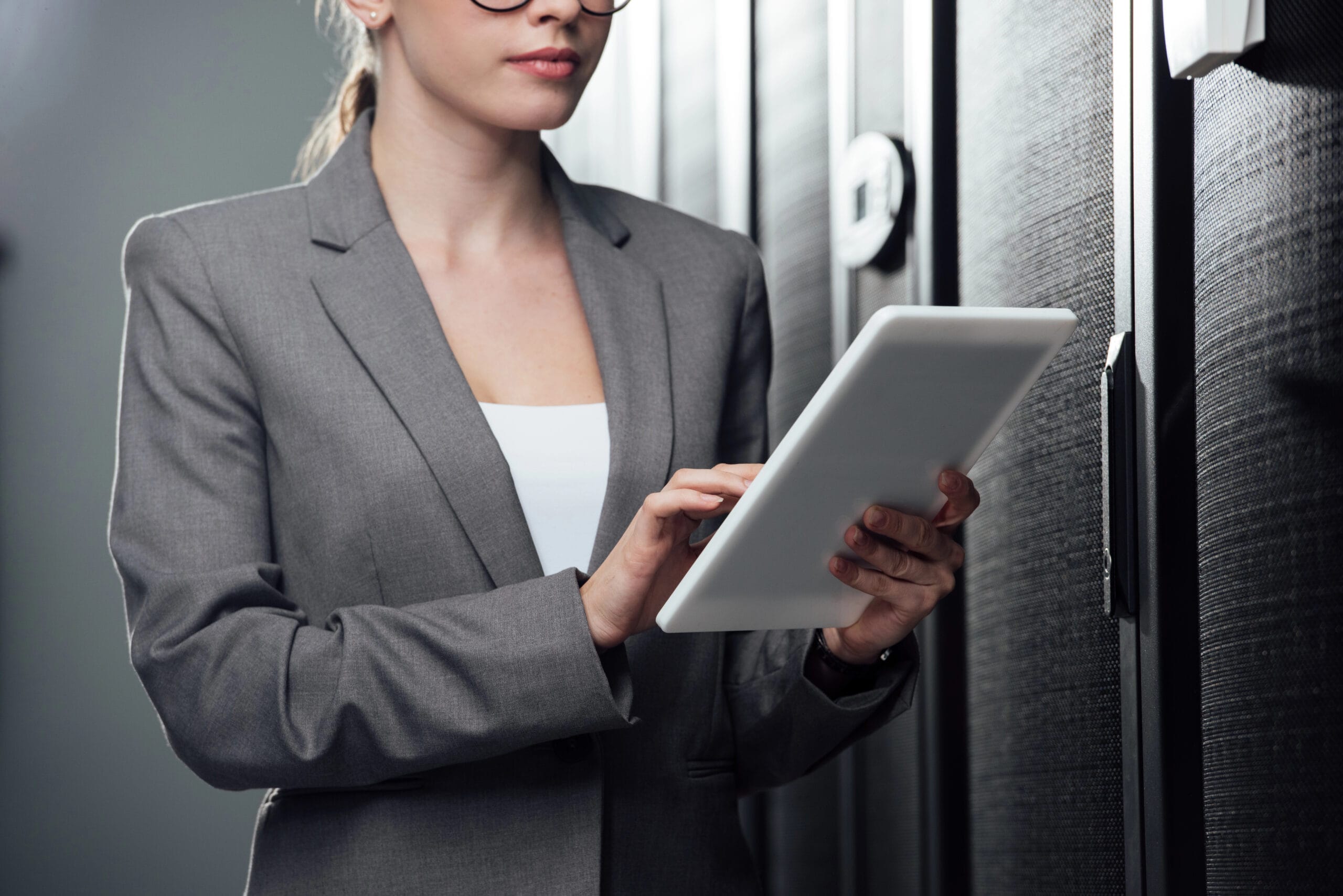 A person in a gray blazer efficiently manages database cleanup using a tablet, standing poised in front of black server cabinets.