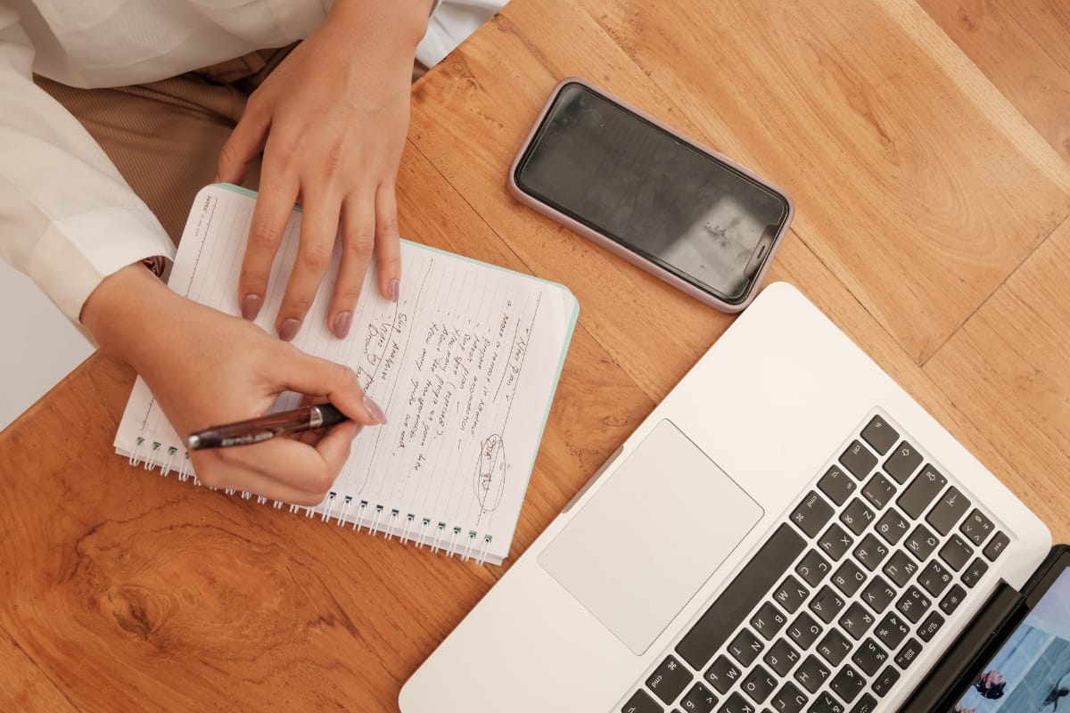 A person engages in content creation, writing notes in a notebook on a wooden desk, with a smartphone and open laptop beside them.