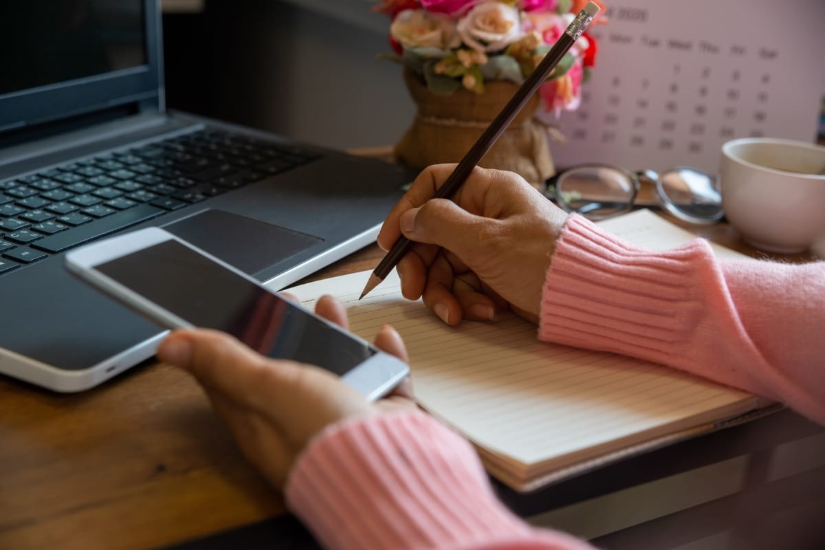 A person sits at a desk, jotting notes in a notebook while holding a smartphone. Nearby, a laptop displays a WordPress backup screen. A fragrant flower arrangement and steaming coffee cup add warmth to the scene.