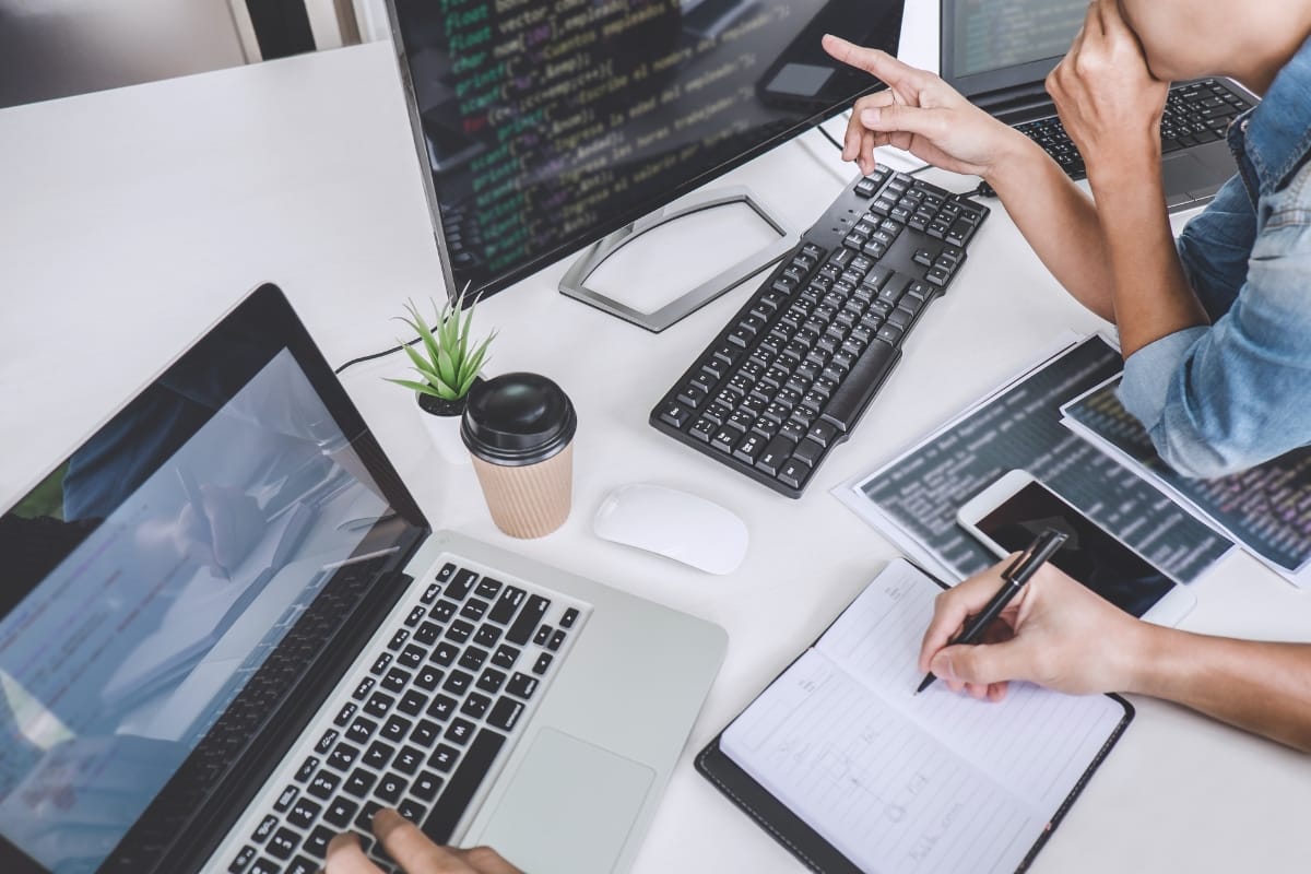 Two people work at a desk with computers and notepads. One points at code on a monitor, discussing WordPress database optimization to improve performance.