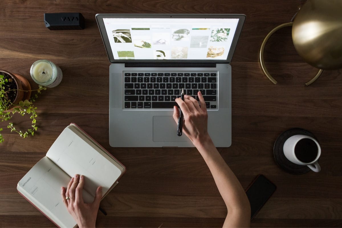 The person is focused on a laptop, delving into image browsing while pondering WordPress database optimization. One hand rests on a notebook as a phone, coffee cup, plant, and candle adorn the wooden table around them.