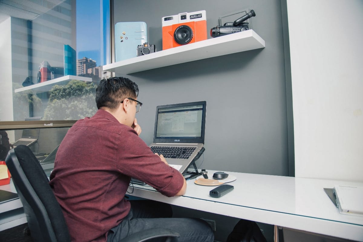 A person in a red shirt is meticulously drafting WordPress posts on a laptop at an office desk. Nearby shelves display a vintage camera and projector, while large windows frame a vibrant cityscape beyond.