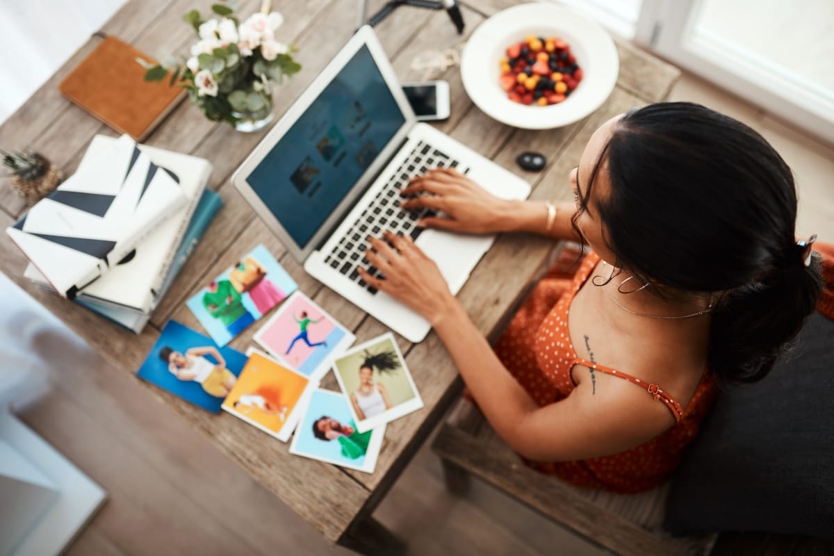 A woman in an orange dress works on a laptop, crafting WordPress posts at a wooden table. Various photos, books, a smartphone, and a bowl of fruit are artfully arranged around her workspace.