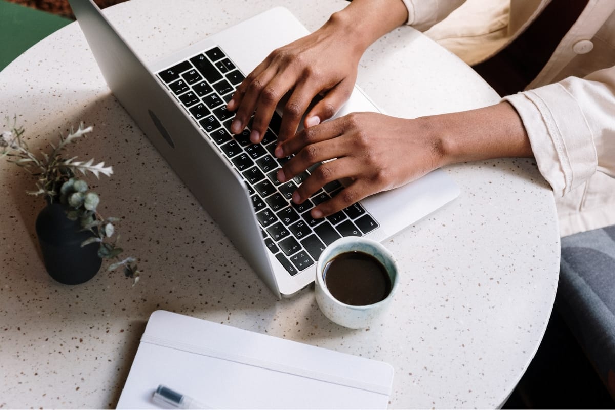 A person diligently typing on a laptop, crafting WordPress posts at a round table. Nearby, a cup of coffee steams gently next to a closed notebook with a pen poised for note-taking.