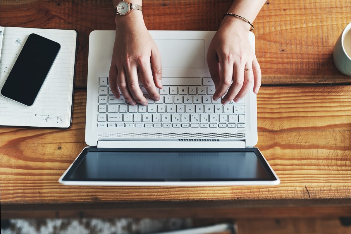 A person types on a laptop at a wooden table, mindful of WordPress security best practices, with a notebook and smartphone nearby.