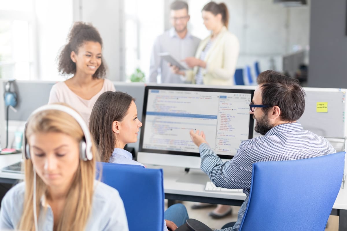 In a modern office setting, people work at computers while a man points at code on a monitor, highlighting key WordPress security best practices. In the background, two colleagues discuss documents, ensuring every aspect of their project aligns with the latest cybersecurity standards.