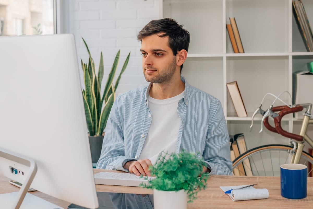 A man sits at a desk working on his computer, focusing intently on WordPress user role management. Nearby, a plant adds a touch of nature, while a notebook and mug suggest productivity. In the background, a bicycle hints at his active lifestyle.