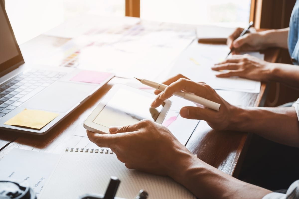 Two people working at a table with a tablet, laptop, and notepads display effective teamwork. One is writing on paper, while the other uses a stylus over the tablet to navigate WordPress user role management tools seamlessly.