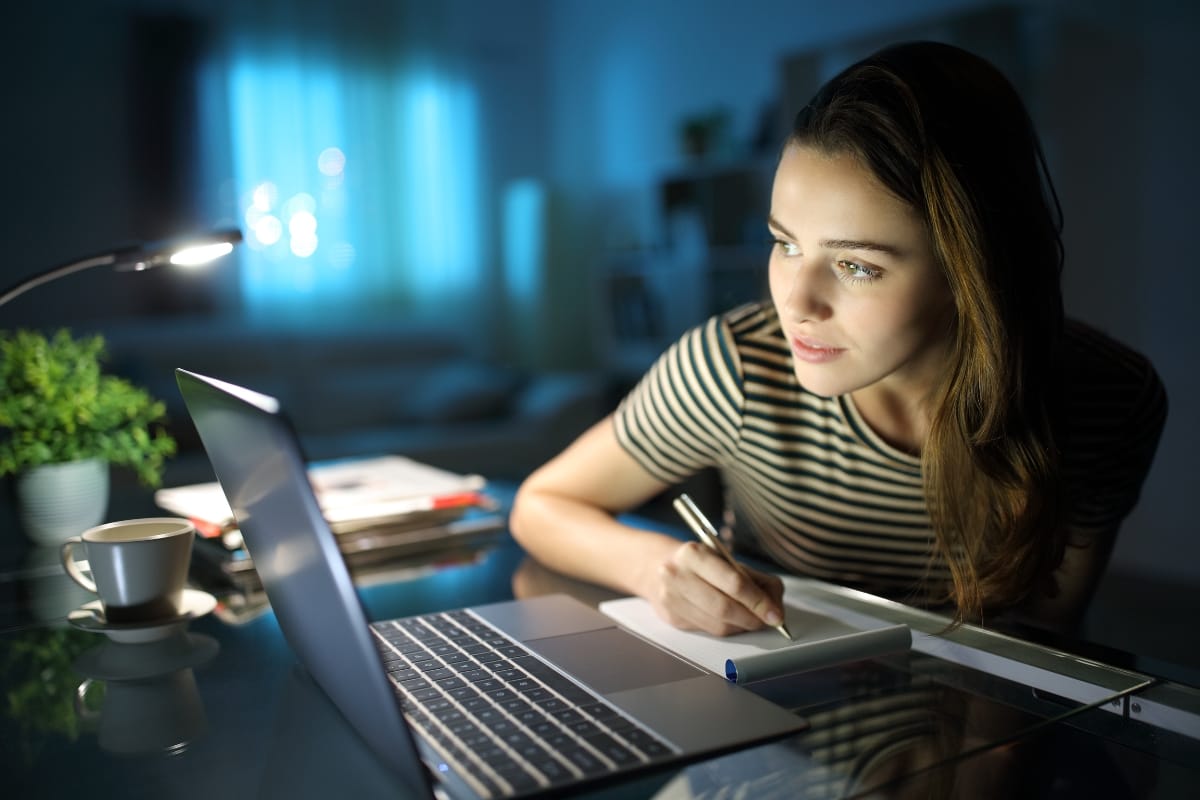 In a dimly lit room, a woman sits at her desk, expertly managing WordPress user roles on her laptop while jotting notes in her notebook. Surrounded by a cup, plant, and books, the warm glow of the lamp casts an inviting ambiance over her focused endeavor.