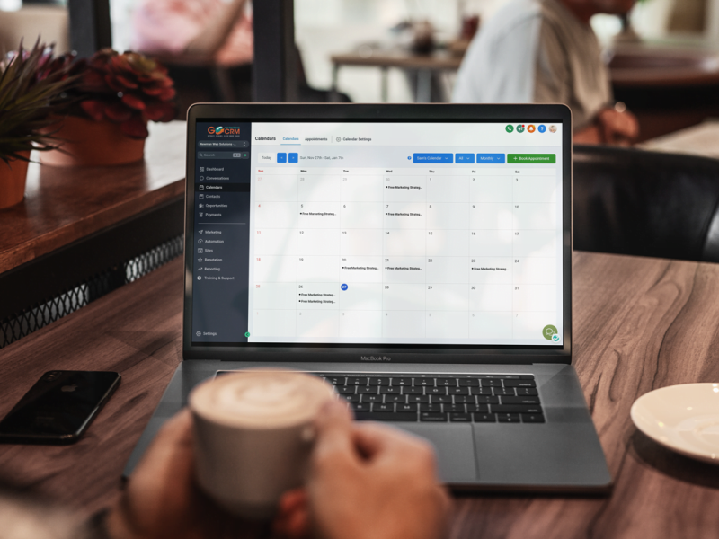 A person holds a cup of coffee while using a laptop displaying a calendar application at a wooden table in a coffee shop.