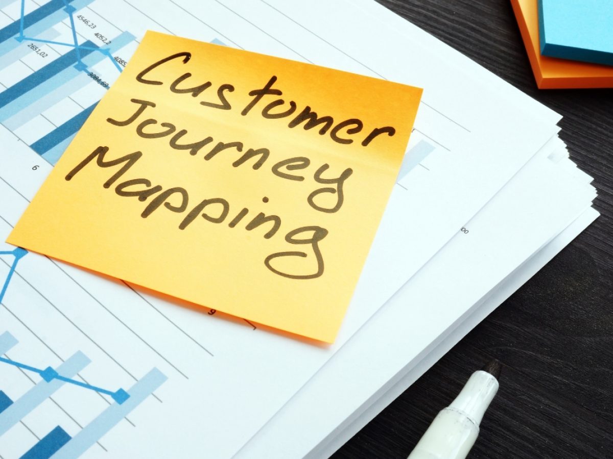 A sticky note with "Customer Journey Mapping" written on it rests atop a stack of charts and graphs, accompanied by a white pen.