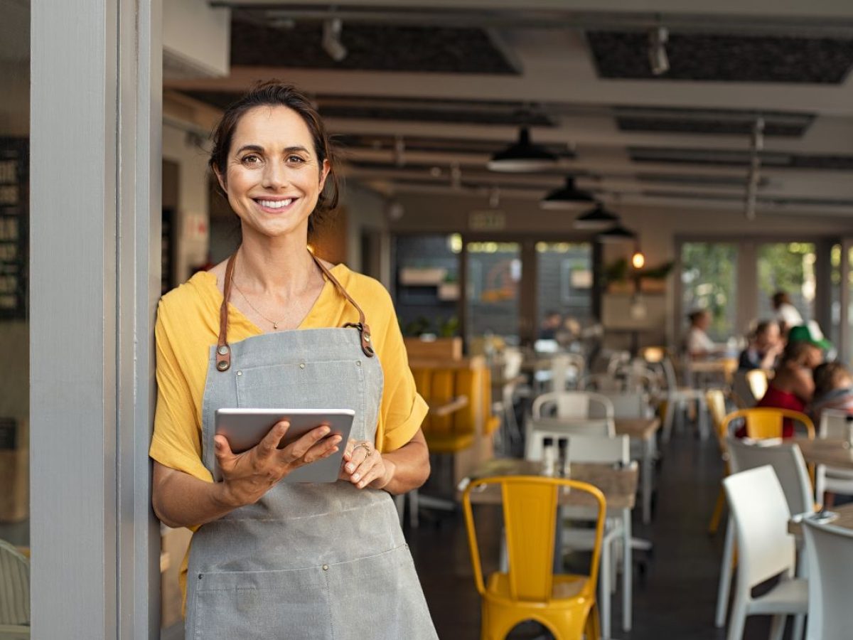 A smiling woman in a yellow shirt and gray apron holds a tablet while standing in a modern, brightly-lit café with customers in the background, seamlessly managing SEO for multiple locations.